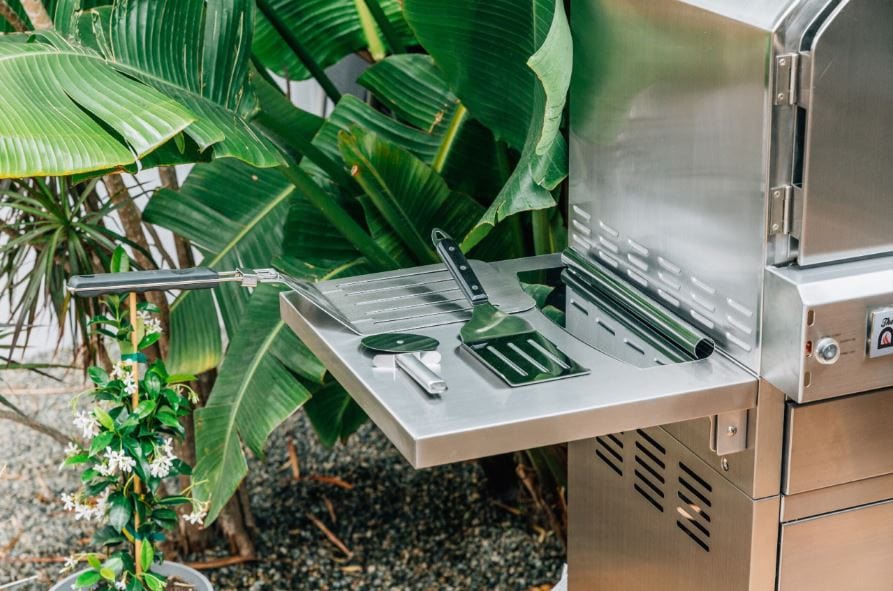 Stainless steel side shelf with grilling tools including a spatula, tongs, and pizza cutter, set against a tropical plant backdrop.