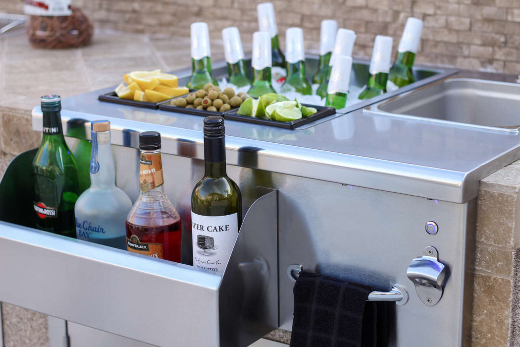 Close-up of an outdoor bar station with a sink, beverage bottles, a towel rack, and a tray holding ice, drinks, lime wedges, and garnishes.