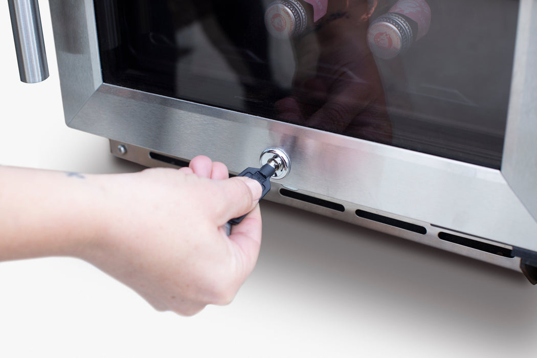 Hand turning a key in the lock of a stainless steel cooler door, with glass panel showing bottles inside and vent slots below.