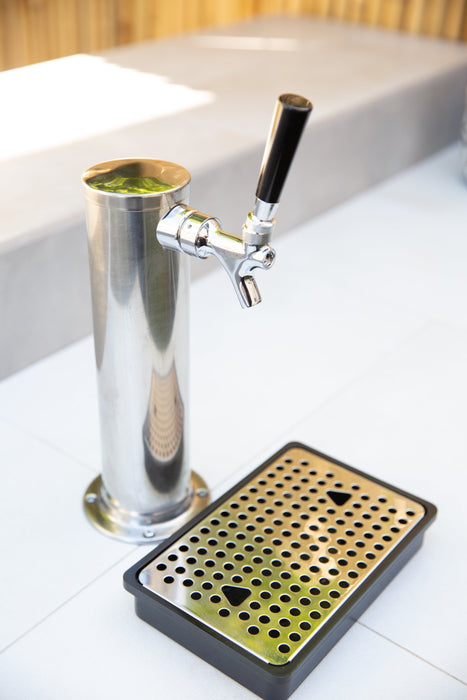 A stainless steel beverage dispenser tap with a black handle, accompanied by a perforated drip tray, set on a white tiled surface.