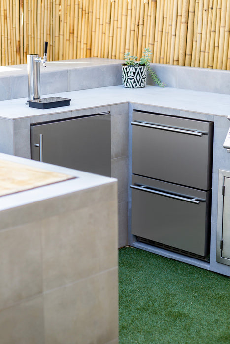 An outdoor kitchen setup featuring a stainless steel beverage dispenser, drawers, cabinets, and a bamboo fence backdrop on artificial grass.