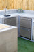 An outdoor kitchen setup featuring a stainless steel beverage dispenser, drawers, cabinets, and a bamboo fence backdrop on artificial grass.