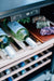 Close-up of a cooler with shelves, white wine bottles, a control panel, and a wooden tray holding assorted cheeses and grapes.