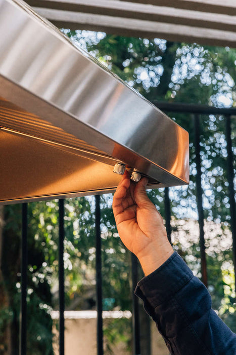 A person adjusting the control knob on a stainless steel outdoor vent hood, with a natural outdoor setting and greenery in the background.