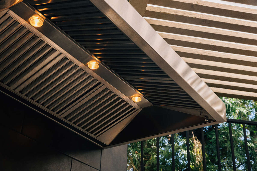 An outdoor vent hood with stainless steel slatted filters and built-in lights, installed under a pergola, with greenery in the background.