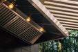 An outdoor vent hood with stainless steel slatted filters and built-in lights, installed under a pergola, with greenery in the background.