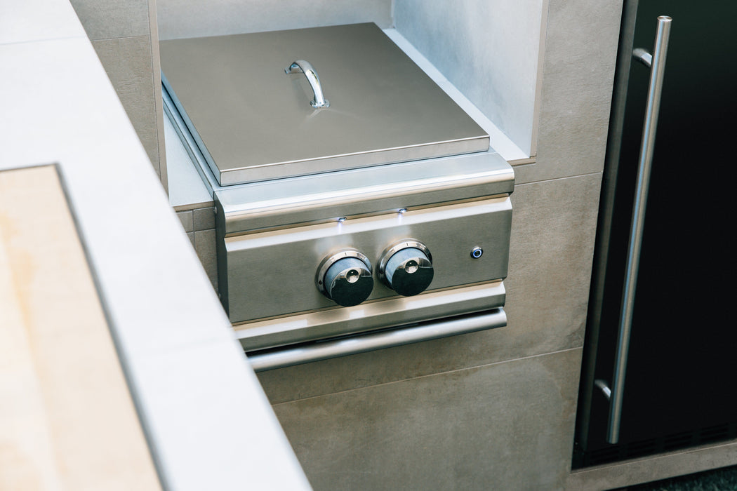 Built-in stainless steel double burner with closed lid and control knobs, integrated into an outdoor kitchen setup with tiled walls.