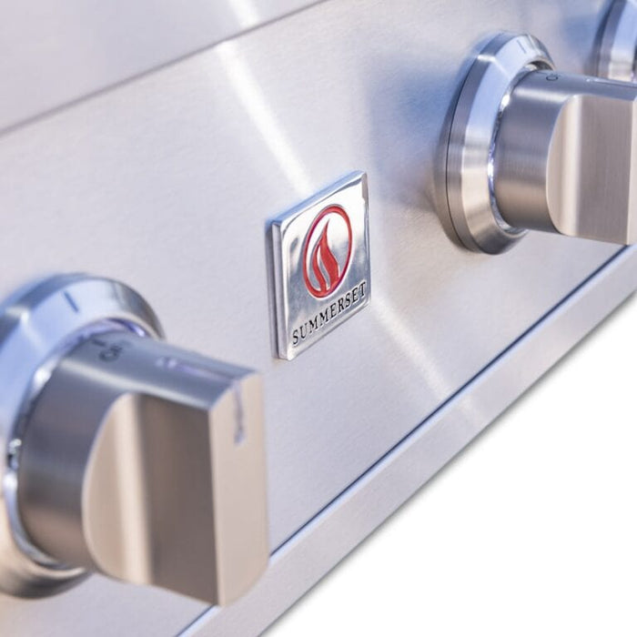 Close-up of stainless steel grill control knobs and logo plate on sleek metallic front panel.