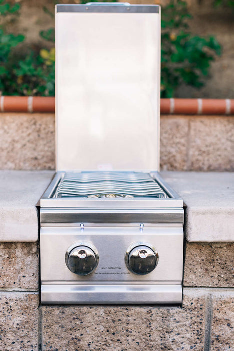 Stainless steel side burner with two knobs and a raised lid, installed in an outdoor kitchen setup against a stone background.