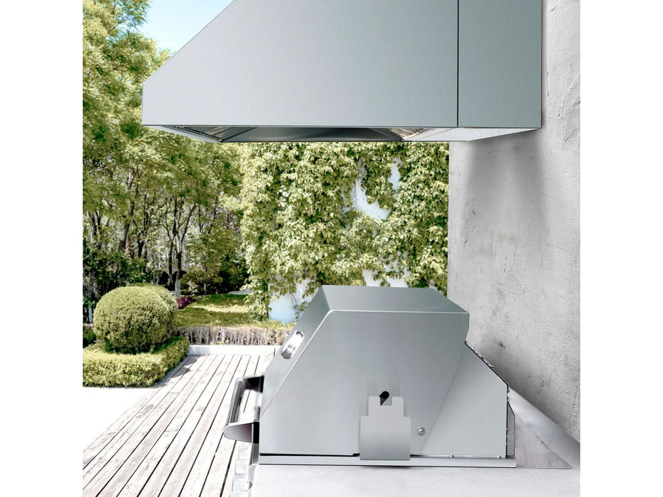 An outdoor cooking area featuring a stainless steel vent hood mounted on a wall above a grill, with lush greenery in the background.