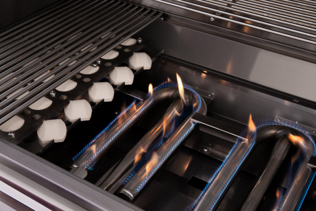 Close-up of a gas grill's interior showing lit burners with blue flames, ceramic briquettes, and stainless steel grates above.