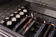 Close-up of a gas grill's interior showing lit burners with blue flames, ceramic briquettes, and stainless steel grates above.