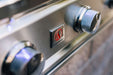 Close-up of a stainless steel grill panel with control knobs and a flame logo in the center, reflecting a polished metallic finish.