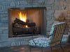 Outdoor stone fireplace with a warm fire, featuring realistic log set, mesh screen, and cozy patio seating with patterned cushions.