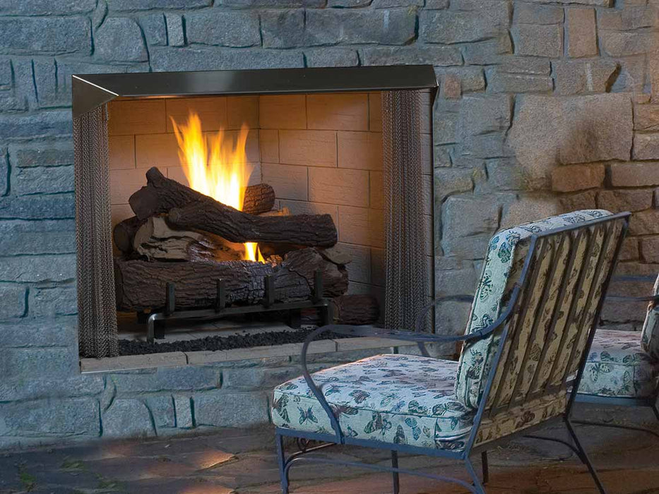 Outdoor stone fireplace with a warm fire, featuring realistic log set, mesh screen, and cozy patio seating with patterned cushions.