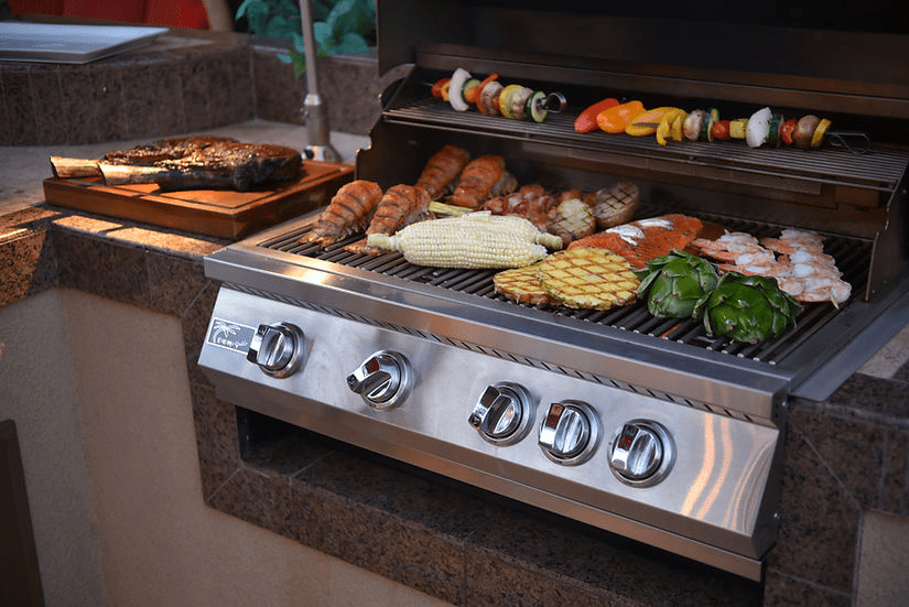 Outdoor built-in gas grill loaded with various foods, including vegetables, shrimp, corn, and skewers, showcasing grilling capabilities and spacious surface