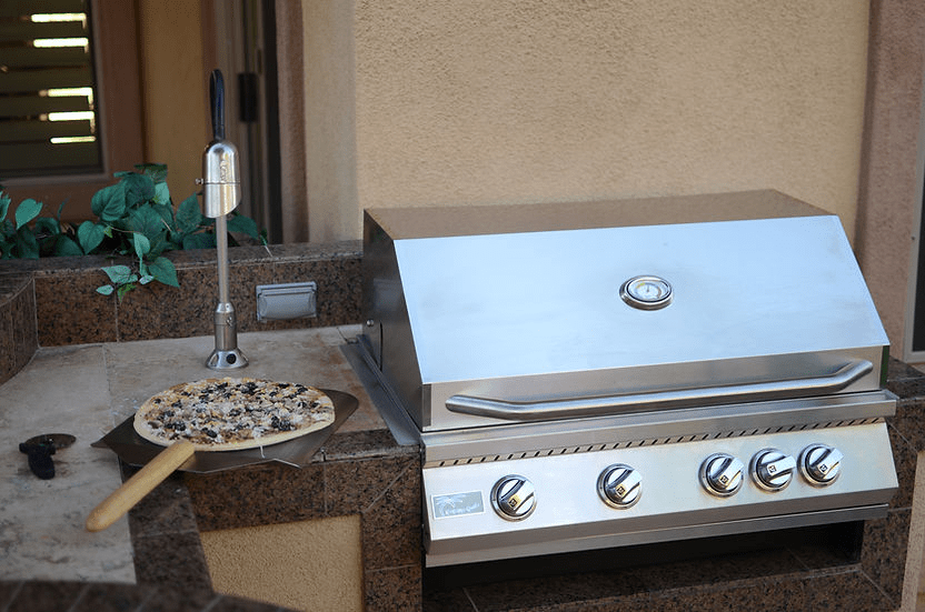 Built-in stainless steel gas grill with closed lid, side prep area holding a pizza on a peel, ideal for outdoor kitchen cooking and entertaining