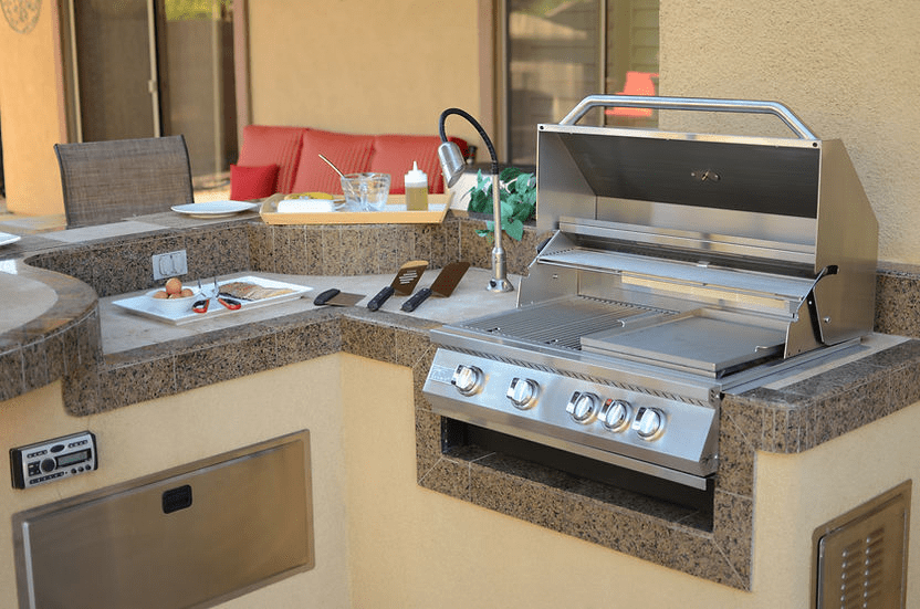 Outdoor kitchen setup with built-in stainless steel gas grill, open lid, side prep area with cooking utensils, and countertop accessories for grilling