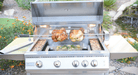 Outdoor stainless steel grill with rotisserie chicken, wood chip trays, and illuminated cooking area, set in a landscaped backyard