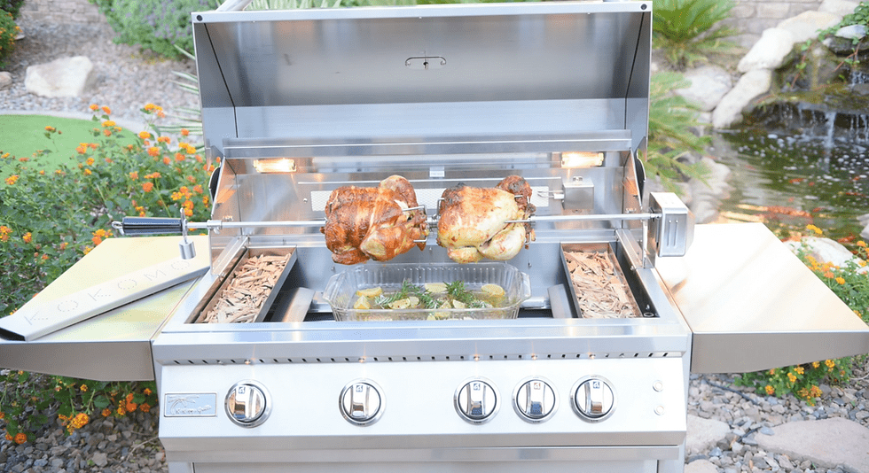 Outdoor stainless steel grill with rotisserie chicken, wood chip trays, and illuminated cooking area, set in a landscaped backyard