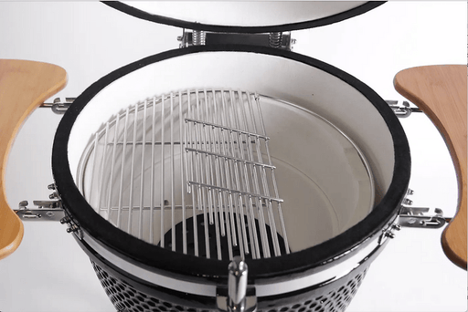 Close up view of open ceramic grill interior showing stainless steel cooking grates, hinged section for fuel addition, and folding wooden side shelves