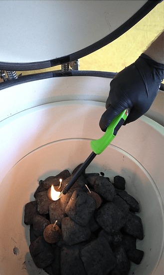 Person lighting charcoal inside a ceramic grill using a long green-handled lighter, with fire starters and with heat-resistant gloves on