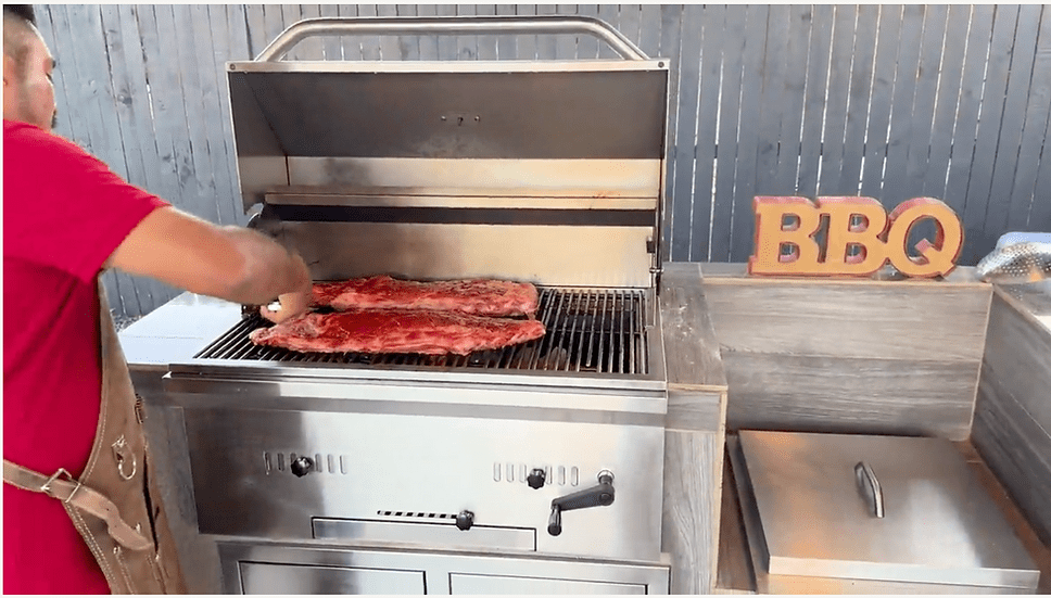 Man grilling ribs on a stainless steel built-in charcoal grill with the lid open, part of a stylish outdoor kitchen setup featuring a BBQ sign