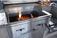 Flames rising from lit charcoal inside a stainless steel built-in grill, showcasing heat control features in a modern outdoor kitchen setup