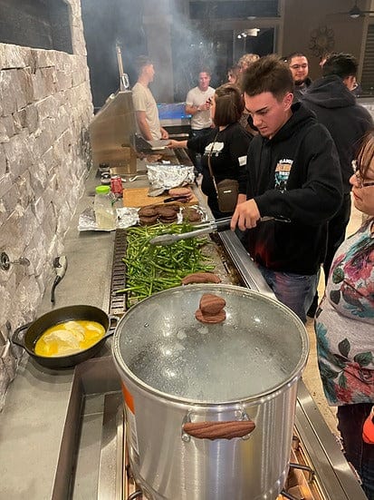 Group of people enjoying an outdoor cooking experience with a built-in grill and griddle, cooking vegetables, burgers, and using a stockpot