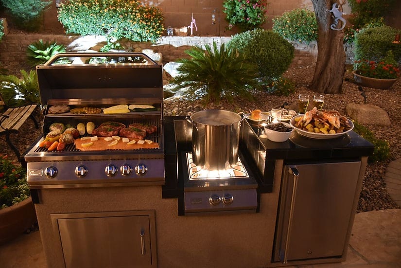 Outdoor kitchen setup at night with a grill cooking meats and vegetables, a burner heating a large stock pot, set in a landscaped backyard