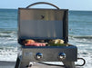 Portable gas grill with open lid grilling steaks, corn, and vegetables by the ocean, showcasing outdoor cooking versatility with a scenic backdrop