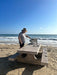 Man grilling on a portable gas grill set on a picnic table by the beach, with waves and a clear blue sky in the background