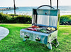 Portable gas grill on green grass, cooking steaks, corn, and vegetables, with a scenic ocean view and pier in the background