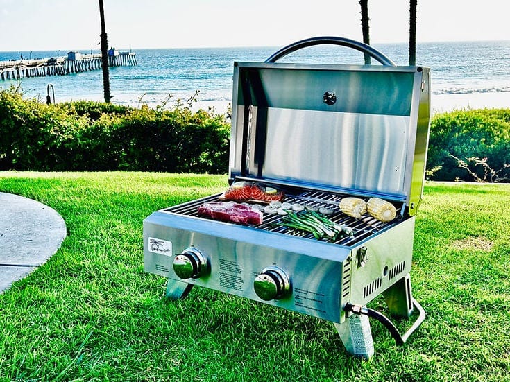 Portable gas grill on green grass, cooking steaks, corn, and vegetables, with a scenic ocean view and pier in the background