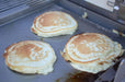 Three golden-brown pancakes cooking on a flat stainless steel griddle, showcasing even heat distribution and a non-stick surface